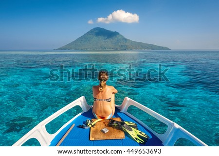 Young woman in swimsuit sit on boat with a mask and flippers looking to a clean sea and volcano Manado Tua. North Sulawesi, Indonesia.