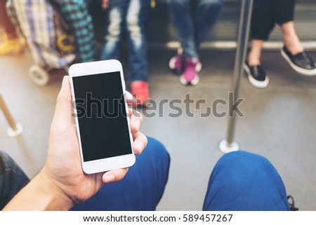 Mockup image of hand holding white mobile phone with blank black screen in subway with many people in background