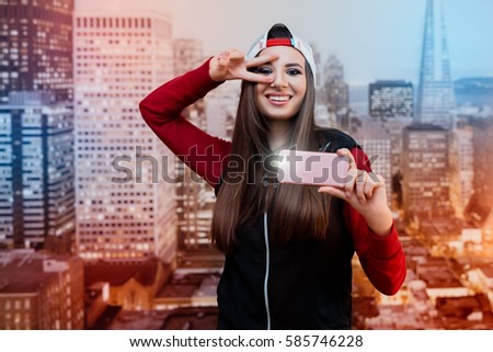 A smiling girl with black eyes. Dressed in black with a red sweater and a cap turned backwards. Itself photographed on a pink smartphone. In the background images of the city at the wallpaper.