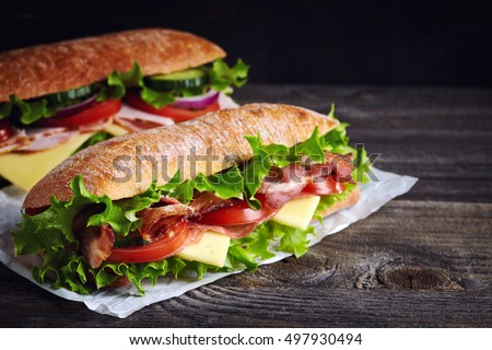 Two fresh submarine sandwiches with ham, cheese, bacon, tomatoes, lettuce, cucumbers and onions on dark wooden background