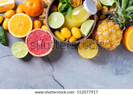 Variety of whole and sliced citrus fruits pineapple, grapefruit, lemon, lime, kumquat, clementine and physalis with mint, bottle of juice in bowl over gray textured background. Top view with space. 