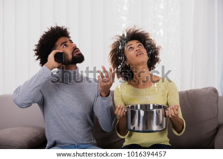 Young Man Calling Plumber While Woman Using Utensil For Collecting Water Leaking From Ceiling