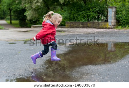 Very cute little girl in red Jacket, blue jeans and rubber boots is jumping over a puddle on a rainy day