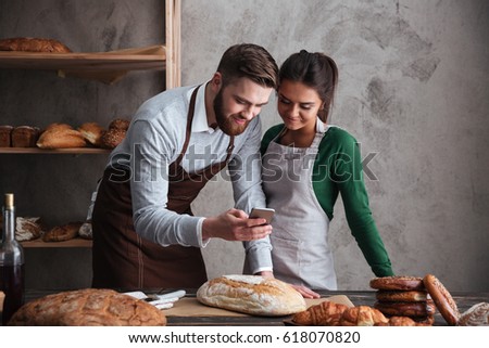 Image of happy loving couple bakers standing near bread and looking at mobile phone.