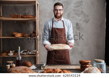 Image of cheerful young man baker standing at bakery holding bread. Looking at camera.