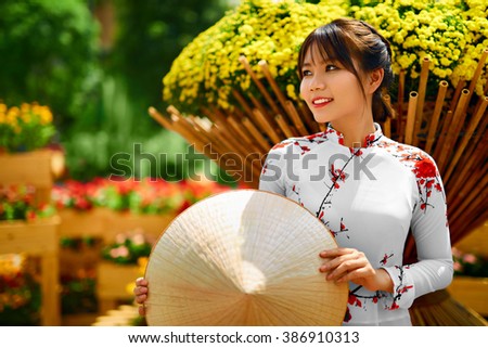 Culture Of Asia. Beautiful Happy Smiling Young Asian Woman Wearing Traditional White Ao Dai Dress ( Clothing ), Vietnamese Conical Hat ( Non La ) Leaf Hat In Flower Garden In Ho Chi Minh City, Vietnam