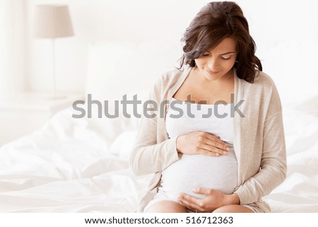 pregnancy, rest, people and expectation concept - happy pregnant woman sitting on bed and touching her belly at home