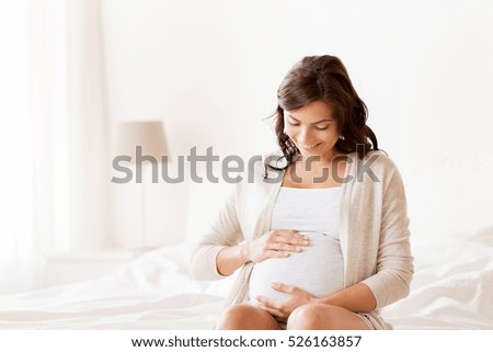 pregnancy, rest, people and expectation concept - happy pregnant woman sitting on bed and touching her belly at home