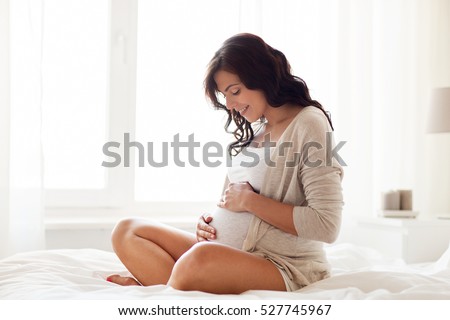 pregnancy, rest, people and expectation concept - happy pregnant woman sitting on bed and touching her belly at home