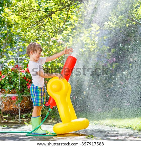 Active happy little kid boy playing and splashing with a garden hose on hot and sunny summer day. Child having fun with sprinkler outdoors. Funny outdoors leisure wth water for children.