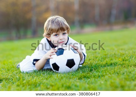 Adorable little kid boy having fun with playing soccer with football on field, outdoors. In white German national uniform. Happy child about european championship