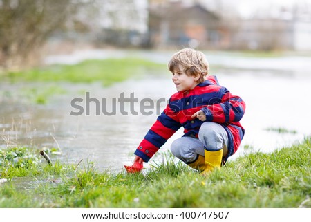 Beautiful little kid boy in rain boots playing with paper boat by a puddle on warm spring day. Active leisure for children. Child having fun outdoors.
