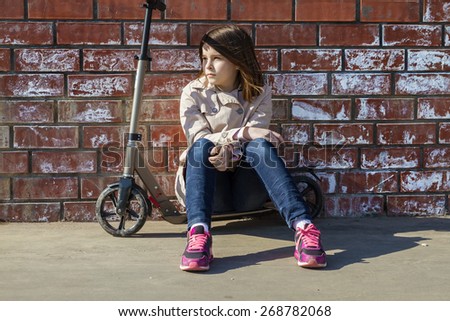 Cute little girl in a light coat and jeans sitting on a scooter in a brick wall in the city