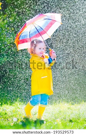 Funny cute curly toddler girl wearing yellow waterproof coat and boots holding colorful umbrella playing in the garden by rain and sun weather on a warm autumn or summer day