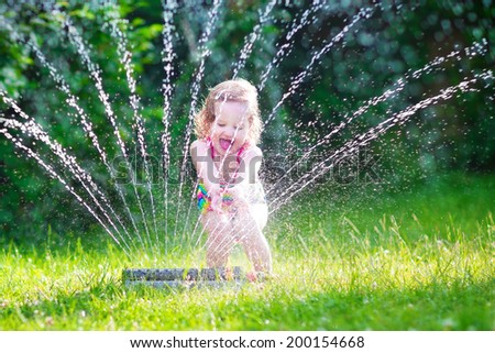 Funny laughing little girl in a colorful swimming suit running though garden sprinkler playing with water splashes having fun in the backyard on a sunny hot summer vacation day