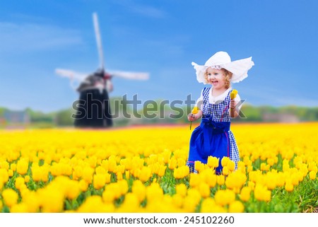 Adorable curly toddler girl wearing Dutch traditional national costume dress and hat playing in a field of blooming tulips next to a windmill in Amsterdam region, Holland, Netherlands