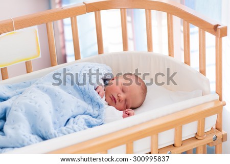 Newborn baby in hospital room. New born child in wooden co-sleeper crib. Infant sleeping in bedside bassinet. Safe co-sleeping in a bed side cot. Little boy taking a nap under knitted blanket.