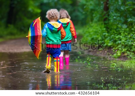 Little boy and girl play in rainy summer park. Children with colorful rainbow umbrella, waterproof boots jump in puddle and mud in the rain. Kids walk in autumn shower. Outdoor fun by any weather