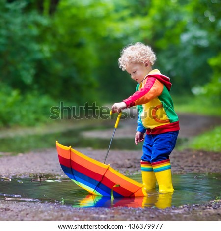 Little boy playing in rainy summer park. Child with rainbow umbrella, waterproof coat and boots jumping in puddle and mud in the rain. Kid walking in autumn shower. Outdoor fun by any weather