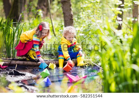 Children play with colorful paper boats in a small river on a sunny spring day. Kids playing exploring the nature. Brother, sister having fun at a forest stream. Boy and girl with toy boat and ship