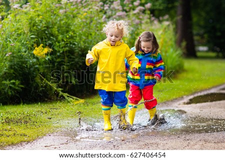 Little boy and girl play in rainy summer park. Children with colorful rainbow jacket and waterproof boots jump in puddle and mud in the rain. Kids walk in autumn shower. Outdoor fun by any weather