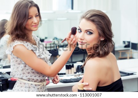 Make-up artist applying powder with a brush on model's cheeks, selective focus on model looking at camera