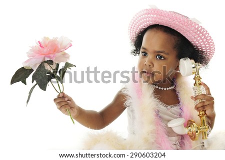 Close-up of an adorable two year old diva in pearls, a pink hat, boas and holding a flower while talking on a fancy French phone. On a white background.