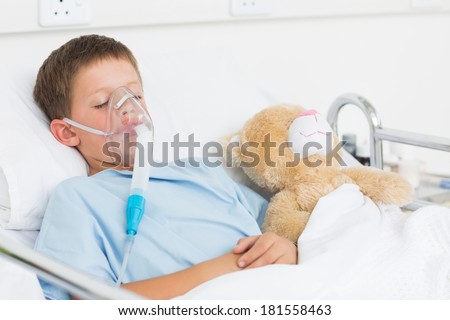 Menino doente usando máscara de oxigênio dormindo ao lado de ursinho de pelúcia na cama do hospital