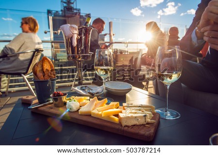 People eating cheese and drinking wine at rooftop restaurant at sunset time. Restaurant table served with cheese plate, bread and white vine full of visitors.