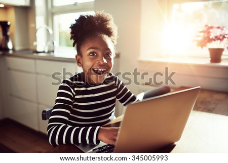 Black girl sitting playing on a laptop computer at home looking at the camera with a joyful expression of amazement and wonder