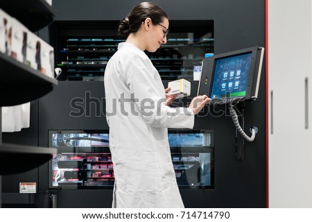 Low-angle rear view of an experienced female pharmacist using a computer while managing the drug stock in a contemporary pharmacy with modern technology