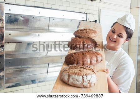 Baker woman presenting bread on board in bakery looking proudly into the camera