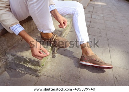 Man in white trousers sitting on the steps in the city and tying the laces on the shoes. Men's summer fashion, stylish men's shoes. Brown Perforated suede summer shoes with laces