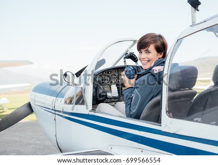 Smiling female pilot in the light aircraft cockpit, she is holding aviator headset and looking at camera