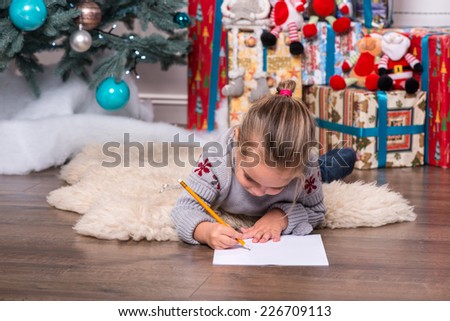 Selective focus on the pretty little fair-haired girl wearing warm sweater and jeans lying on the floor near the Christmas tree writing very diligently something