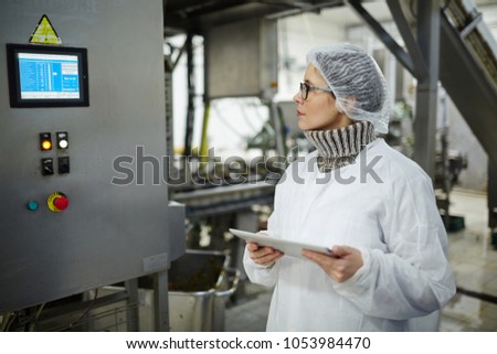Staff of food factory in protective uniform looking at display with regullation information on wall of industrial machine