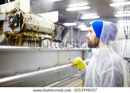 Staff in protective coveralls looking at processing machine mixing seaweed salad before canning