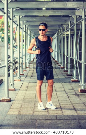 Man summer casual street fashion in New York. Wearing black tank top, shorts, white sneakers, cap worn backward, sunglasses, shoulder carrying bag, a guy stands on sidewalk bridge. Filtered effect.