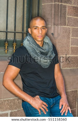 Man Street Casual Fashion in New York. Wearing black T shirt, blue jeans, long patterned scarf around neck, Mixed Race French guy with shaved head stands against vintage wall.