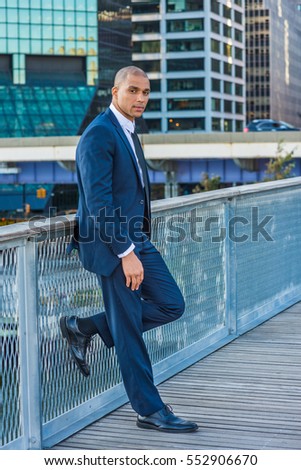 European Businessman with shaved head traveling, working in New York, wearing blue suit, white shirt, black tie, standing by fence in business district, taking work break.