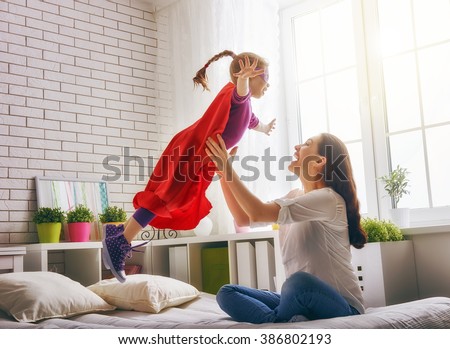 Mother and her child girl playing together. Girl in an Superman's costume. The child having fun and jumping on the bed.