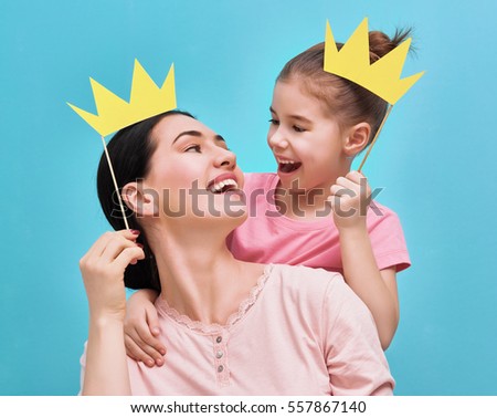 Funny family on a background of bright blue wall. Mother and her daughter girl with a paper accessories. Mom and child are holding paper crown on stick.