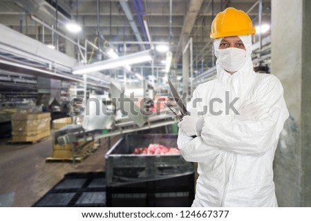 Industrial butcher posing with two filleting knives, wearing protective and hygienic clothing, such as a white suit, mask and a yellow hard hat, in front of a large animal processing plant