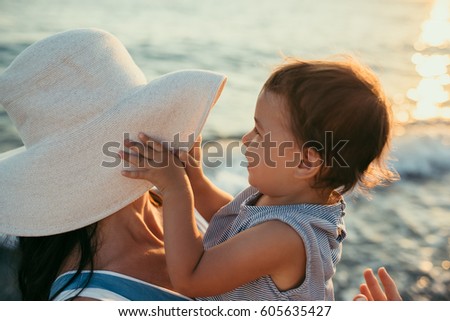 Top view close up of young mother playing with daughter with white hat at the sea background. Family vacation. Travel. Female play with cheerful toddler expression. Caucasian female with baby at ocean