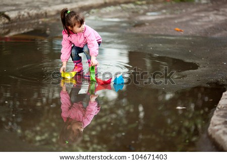 little girl, wearing a pink jacket, playing with colorful paper ship, in the puddle