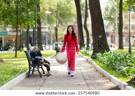 Elegant young Vietnamese woman wearing a traditional red dress walking through a public park past a bench with a person sitting on it