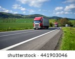 Three trucks driving on asphalt road between green fields in the countryside. Wooded mountains in the background. Sunny day with blue skies and white clouds.