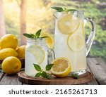 Glass and pitcher of lemonade with lemon slices and mint on an old wooden table with rural summer background