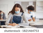 Small photo of High school student taking notes while wearing face mask due to coronavirus emergency. Young woman sitting in class with their classmates and wearing surgical mask due to Covid-19 pandemic.