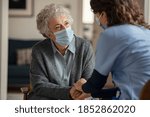Small photo of Elderly woman talking with a doctor while holding hands at home and wearing face protective mask. Worried senior woman talking to her general practitioner visiting her at home during virus epidemic.